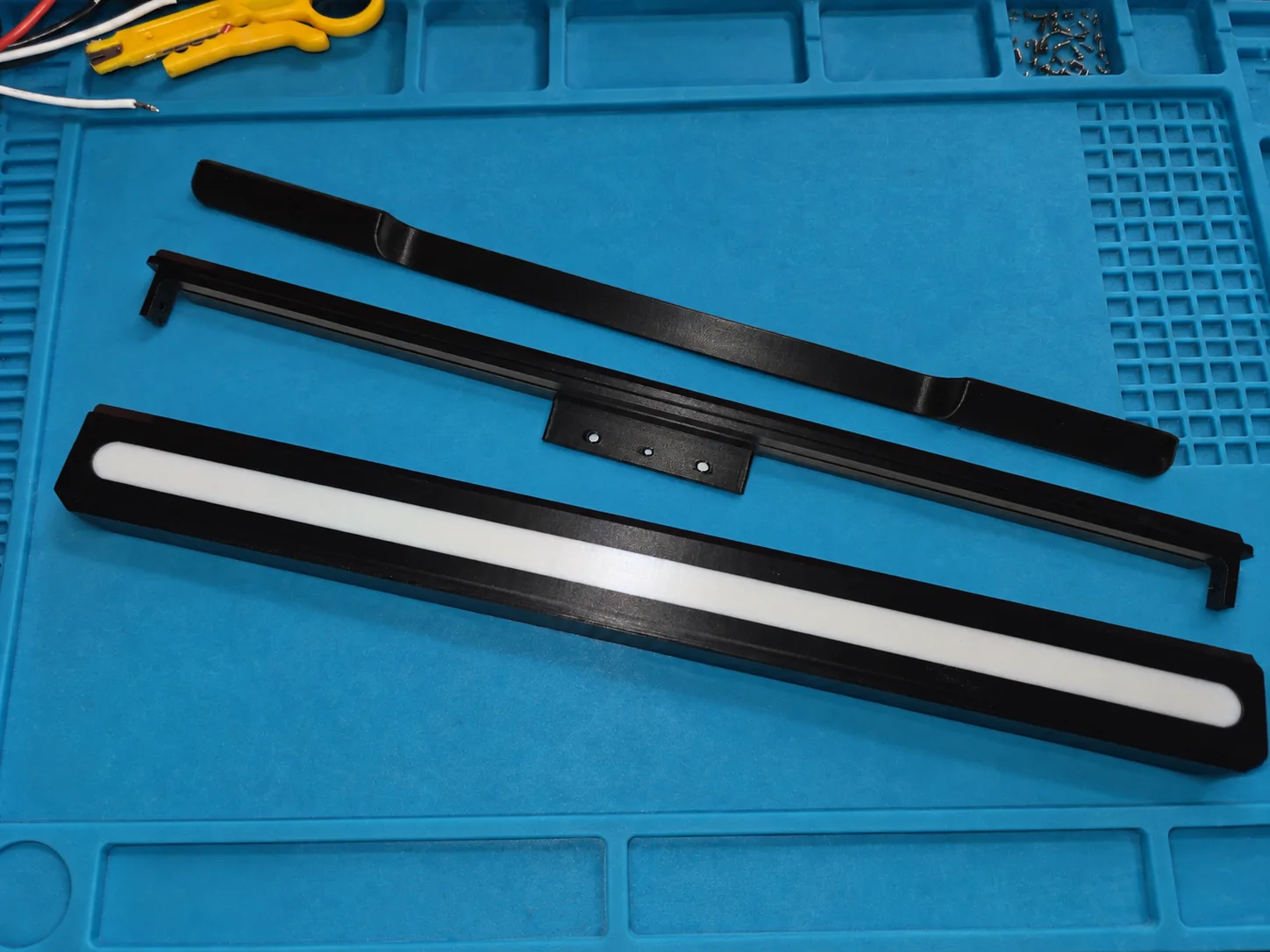 Drei schwarze 3D-gedruckte Teile auf einer blauen Werkstattmatte: ein Gehäuse mit weißem Streifen und zwei Halteteile.