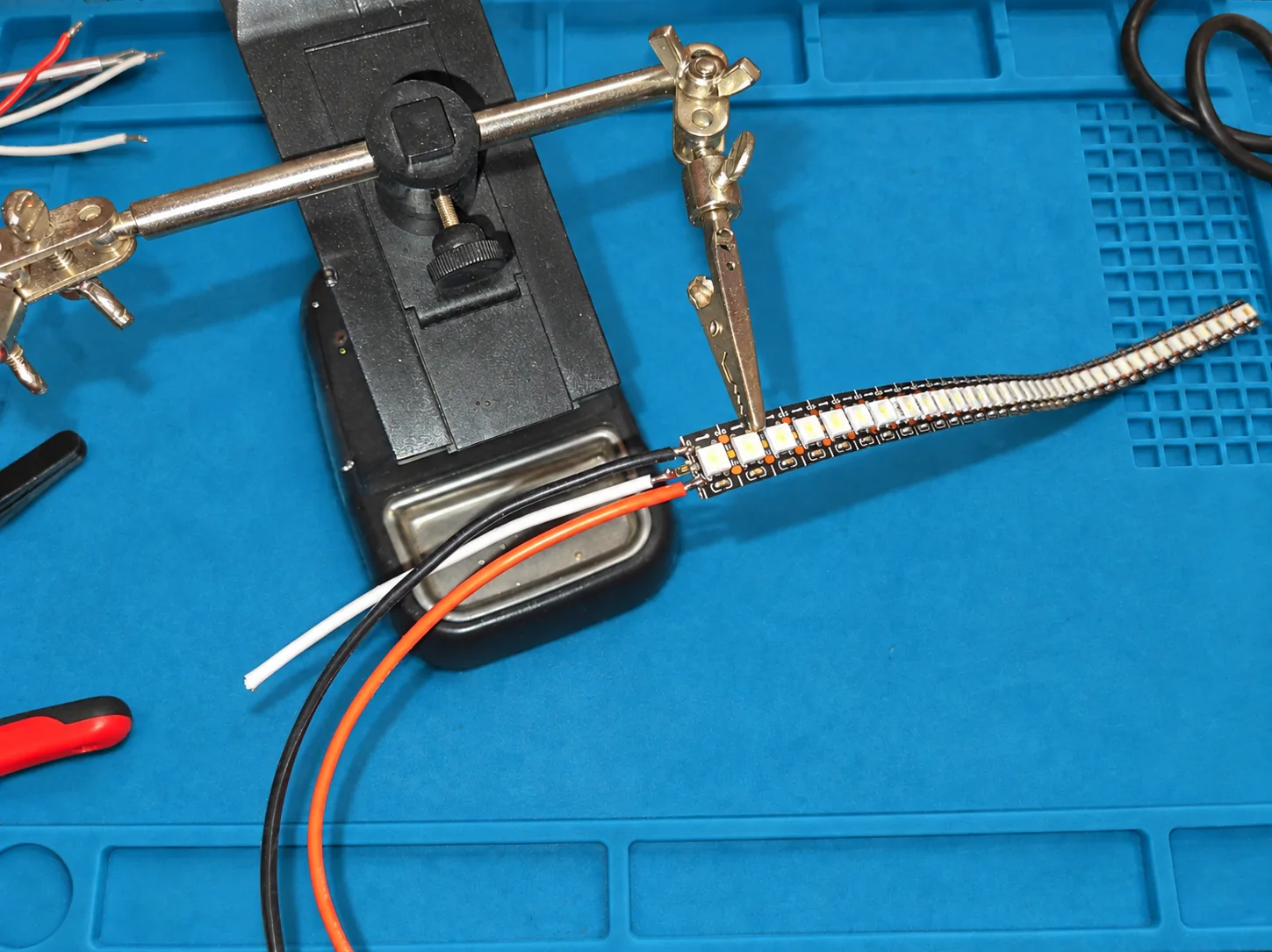 Ein LED-Streifen in einer Haltehilfe, daran drei angelötete Kabel in Rot, Schwarz und Weiß.